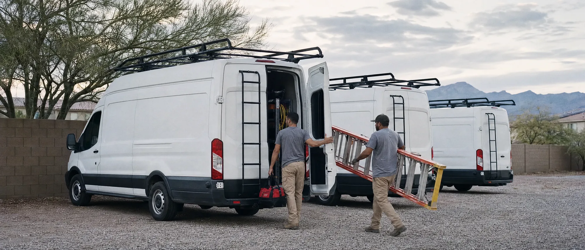 Three white service vans with ladder racks in a Phoenix contractor's yard at dawn